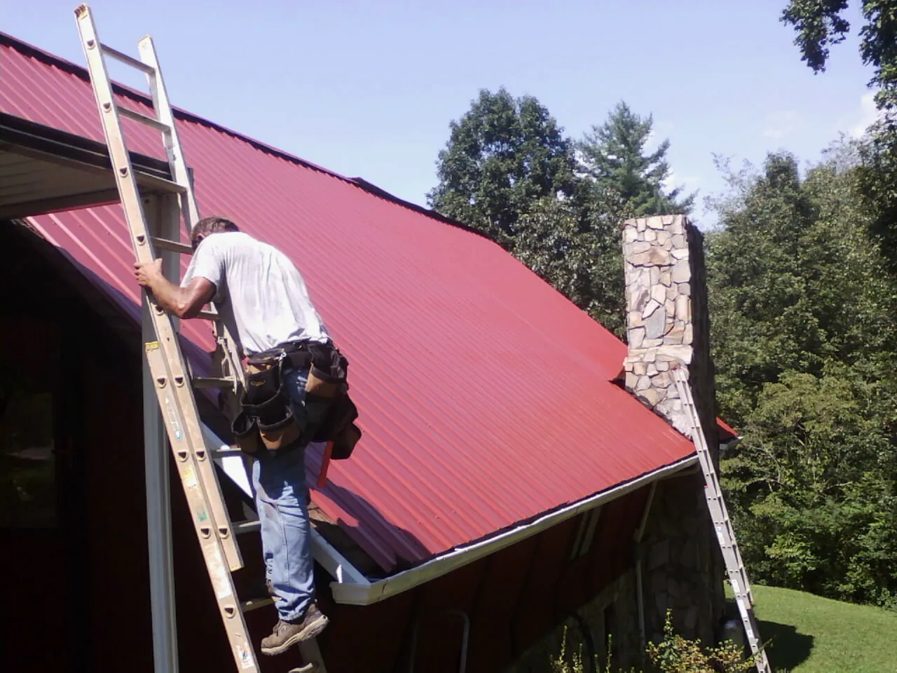 Roofer on a ladder beside a red metal roof and stone chimney — Soffit & Fascia Repair by Roofing Brothers in Strathmere